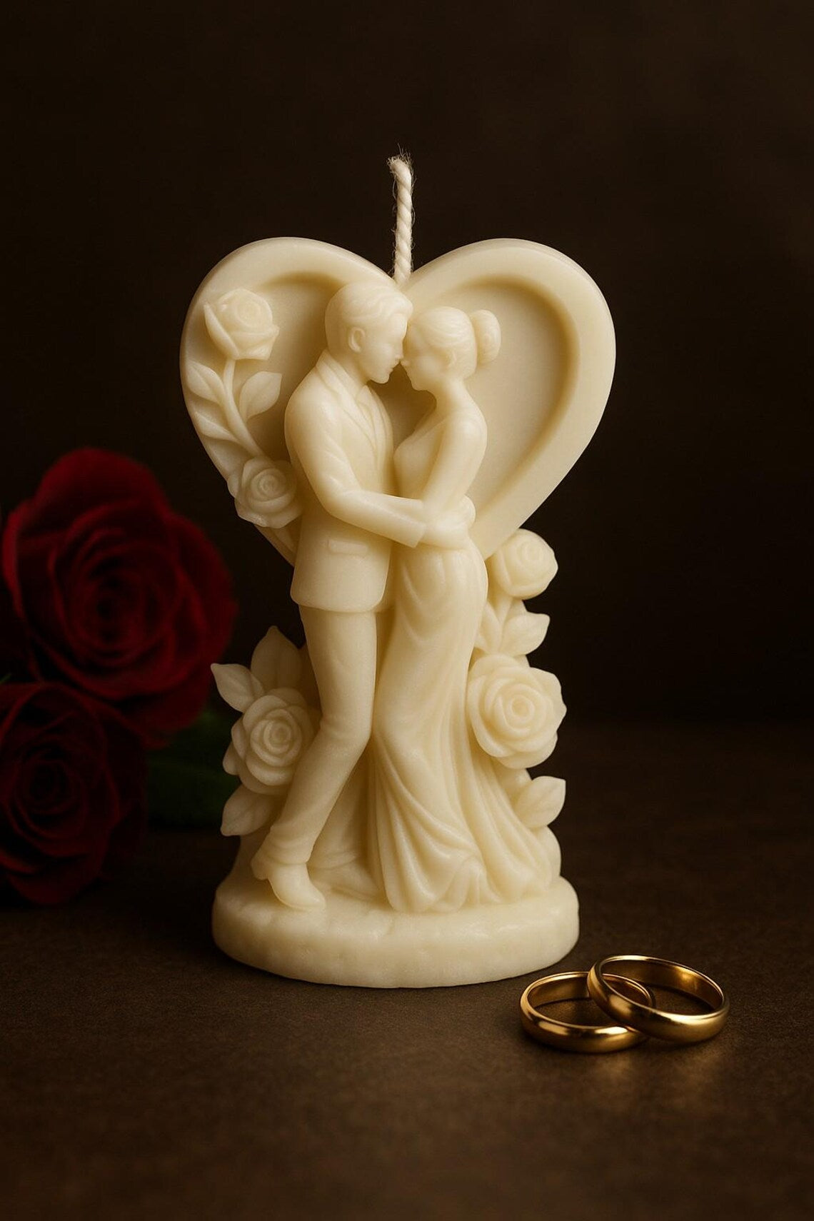 Cremefarbene Kerze in Herzform mit liebevoll gestalteter Paar-Figur, die sich umarmt, verziert mit Rosenmotiven; im Vordergrund liegen zwei goldene Ringe, dunkler, eleganter Hintergrund.