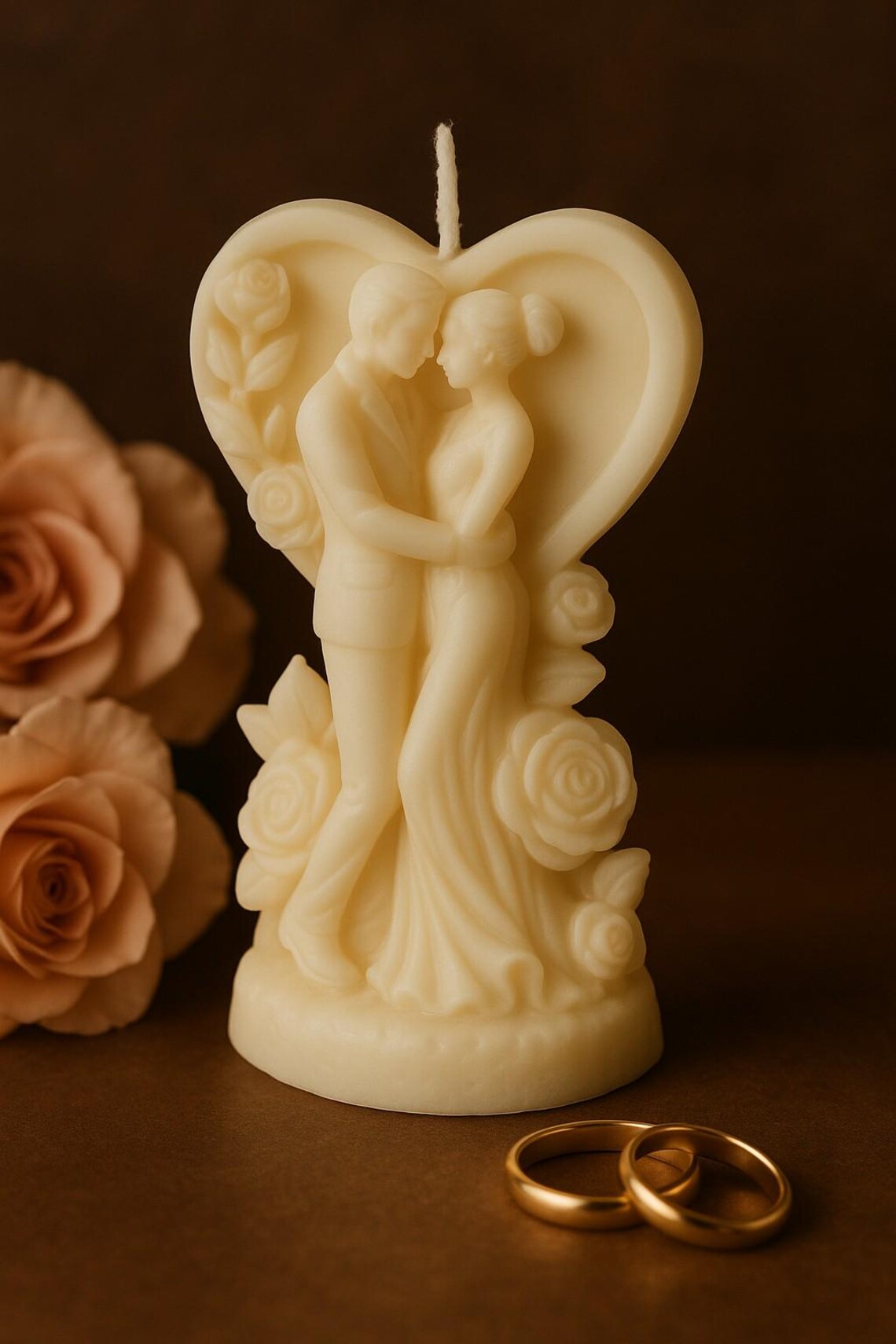 Cremefarbene Kerze in Herzform mit detailreicher Paar-Figur, die sich liebevoll umarmt, umgeben von Rosenmotiven; im Vordergrund liegen zwei goldene Ringe, warme romantische Lichtstimmung.