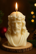 Brennende, cremefarbene Kerze in Form eines Jesuskopfes mit Dornenkrone, auf rundem Holzsockel; weihnachtliche Dekoration mit Tannenzweigen, Kugeln und warmen Lichtern im Hintergrund.
