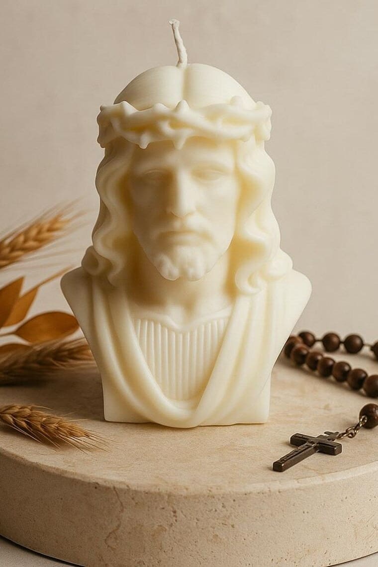 Cremefarbene Kerze in Form eines Jesuskopfes mit Dornenkrone, unangezündet, auf rundem Steinsockel arrangiert mit Rosenkranz und getrockneten Ähren vor hellem, ruhigem Hintergrund.